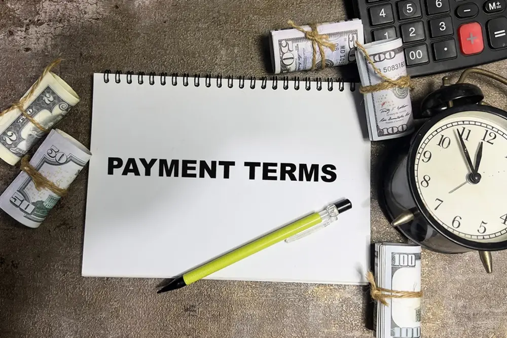 A notebook labeled "Payment Terms" surrounded by cash, pen, and clock—relevant for booking international vacation deals or travel plans from Bangladesh