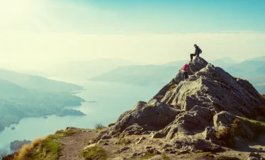 Travelers hiking a rocky mountain peak with scenic lake views, offering a unique adventure experience beyond typical Maldives tour packages from Bangladesh