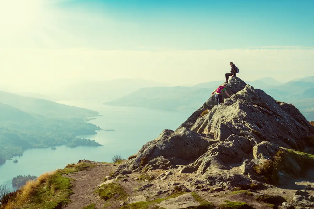 Travelers hiking a rocky mountain peak with scenic lake views, offering a unique adventure experience beyond typical Maldives tour packages from Bangladesh